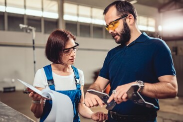 Ein Mann und eine Frau – jeweils mit Arbeitsschutzbrille – unterhalten sich über einen Gegenstand